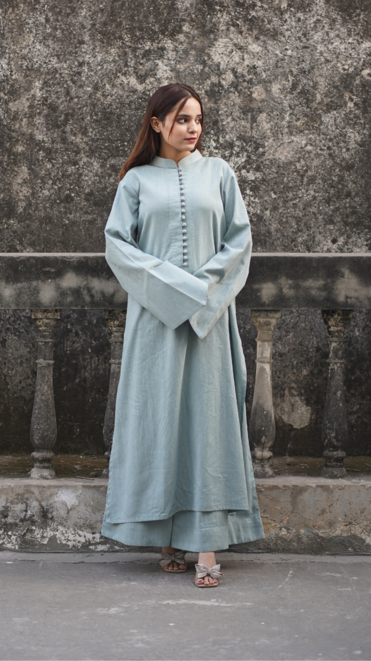 Woman wearing a light blue traditional outfit standing against a textured wall.