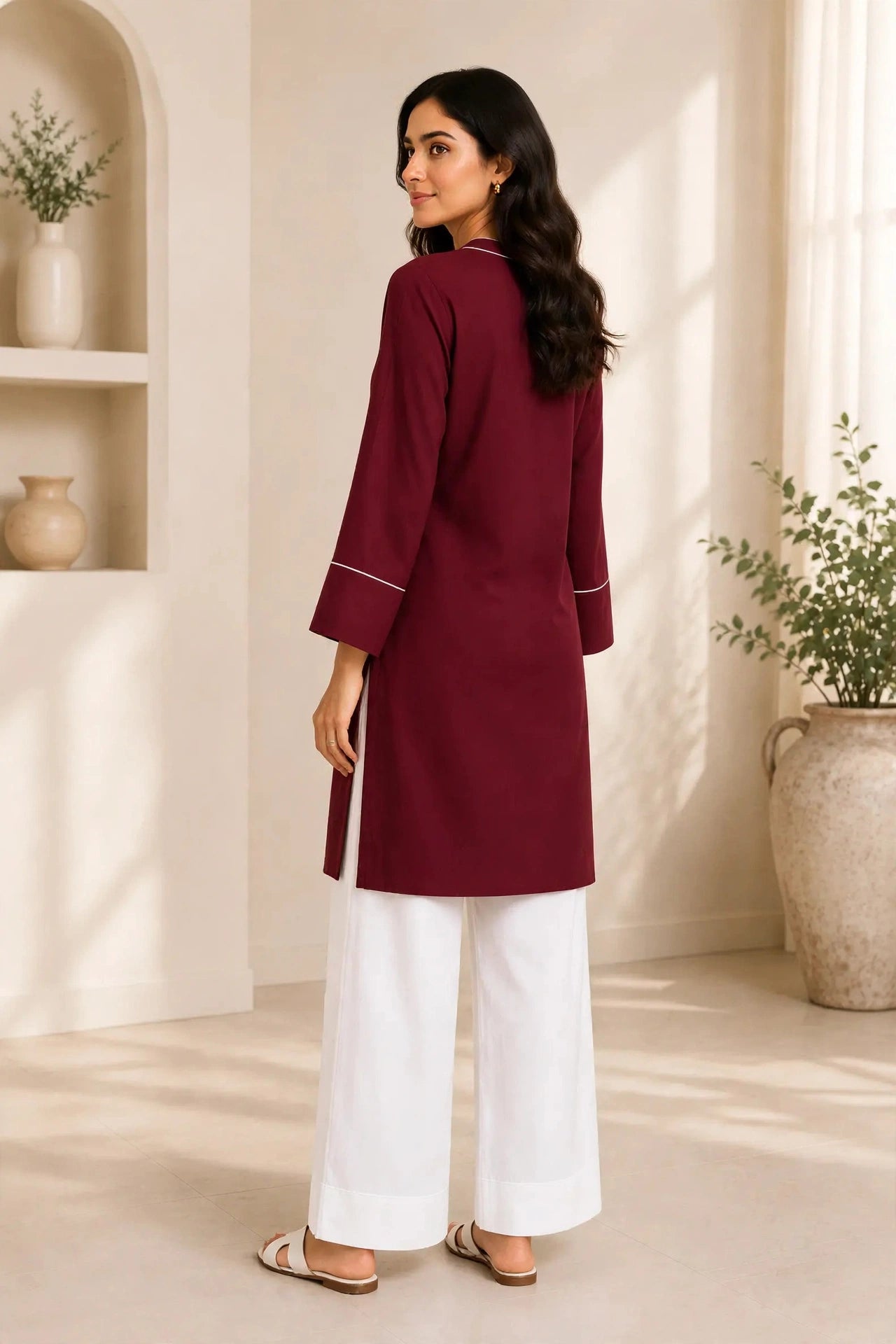 A woman wearing a deep maroon long kurta with white pants, standing in a bright, neutral-toned room with plants and shelves.